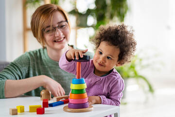 A sitter watches a toddler stacking rings on a stick