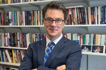 A man wearing glasses stands with his arms folded in front of a large, full bookshelf