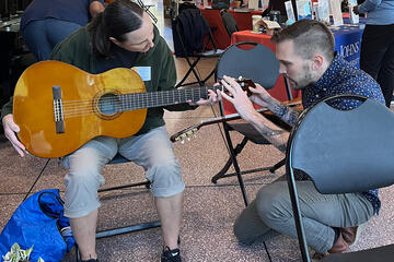 A seated attendee with guitar gets instruction in how to strum along at work.