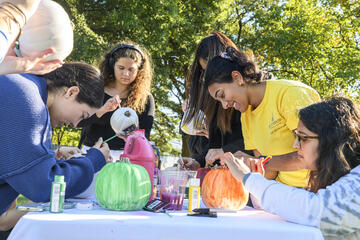 A group of young adults paint pumpkins on an outdoor table.