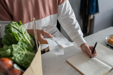 An adult writes down the contents of a receipt in a notebook. Next to them is a paper bag full of produce.