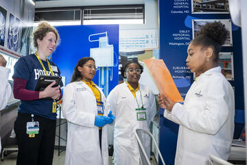 An adult wearing a Hopkins shirt and three children wearing doctor's coats play hospital at Biztown.