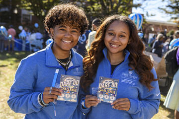 Two college students smile at an outdoor event while holding up small flyers that read 