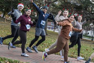 Six runners wearing reindeer antlers or Santa hats wave to the camera as they sprint past. They are wearing numbers taped to their shirts.