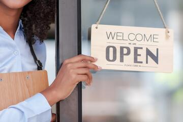 A hand flips a door sign to read "Welcome. We are open. Please come in."