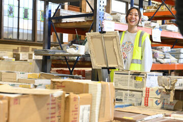 A painter in a yellow safety vest smiles for the camera while standing in a warehouse. In front of them is a canvas and easel, as well as a sea of boxes.