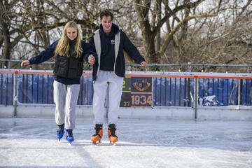 Two adults ice skate hand in hand, looking down at the ice.