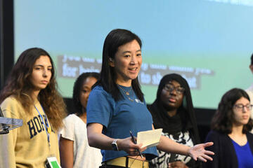 A woman in a blue shirt speaks to a large group