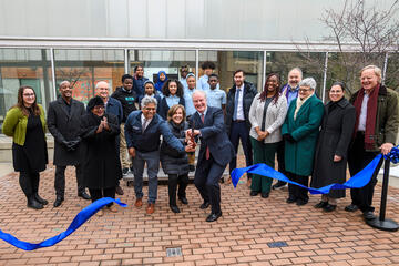 A large group of people participate in a ribbon-cutting ceremony with a blue ribbon