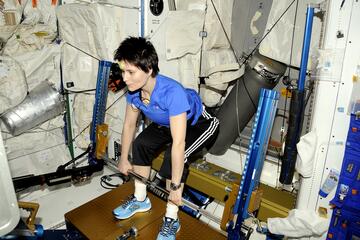 Italian astronaut Samantha Cristoforetti, wearing a blue shirt and black athletic pants, performs a deadlift exercise using the advanced Resistive Exercise Device (aRED) aboard the International Space Station. The aRED machine, equipped with metal bars and vacuum cylinders, provides resistance training to help astronauts maintain muscle and bone strength in microgravity. The surrounding area is filled with white padded walls, equipment, and storage bags, characteristic of the ISS interior.