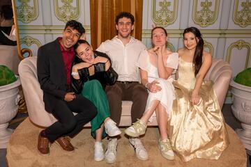 Five college students in formal outfits smile for the camera while sitting on a fancy couch.