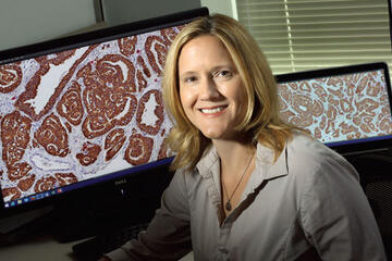 A researcher in a white lab coat sits in front of computer screens showing close-up images of cells