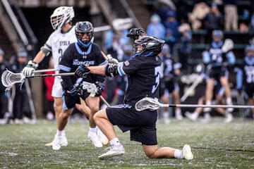 A lacrosse player in a black jersey kneels on one knee to celebrate a goal