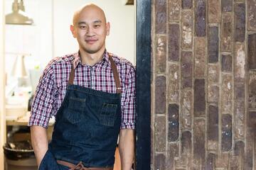 A smiling Tim Ma leans against a wall while wearing a kitchen apron.