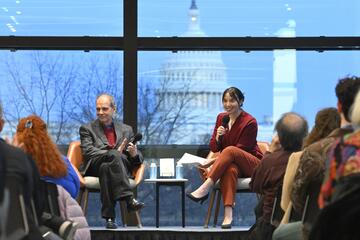 Arturo Casadevall and Emily Kwong sit on a stage in front of a crowd. Behind them, a large window reveals the US Capitol building.