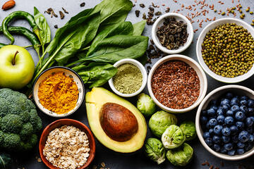 Tabletop covered with superfoods that include avocado, brussel sprouts, blueberries, and seeds.