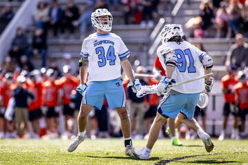 Two men's lacrosse players in white jerseys celebrate a goal