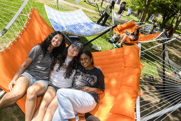 Three young adults smile for the camera while sitting on an outdoor hammock. Behind them are two other hammocks.