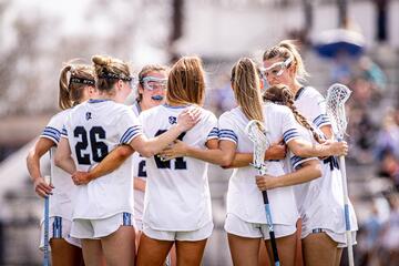 A group of women's lacrosse players in white huddle with their arms around one another