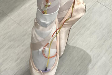 Photo of someone wearing ballet shoes and the PointeSense system, which consists of red and yellow wires running from the wearer's toes to their ankles