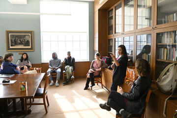 Dora Malech stands at the front of a room lined with books, leading a classroom of high school and college students