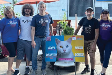 Five volunteers stand in a row next to a sign reading "ADOPT. FOSTER. VOLUNTEER. DONATE."