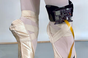 Close-up image of a dancer standing on her toes in ballet pointe shoes; a black control board is wrapped around her right ankle and connected into the toe of the shoe