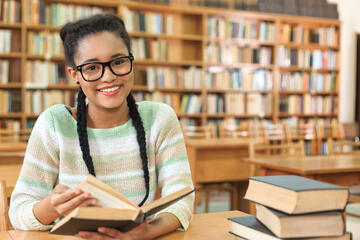 Teenage girl sits at a library table with multiple books 