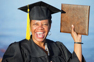 Middle-aged woman in cap and gown holding up her diploma in celebration of receiving it