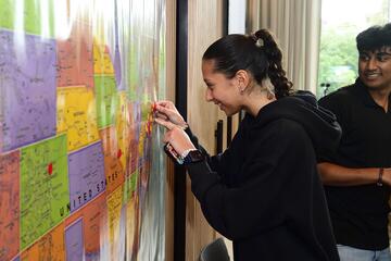 A young adult puts a small, round sticker over New Jersey on a large map of the USA. A few dozen other stickers have already been placed across the map.