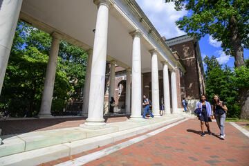 Students walk along a brick pathway with white brick columns on the Homewood campus on a late summer day with blue skies and green trees in the background