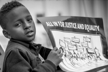 Black-and-white photograph of a young Black boy who is looking at the camera and holding a sign that says "All in for justice and equality"