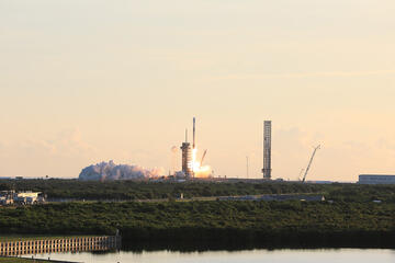 A rocket launches in the distance against a backdrop of a yellow-orange Florida sunrise