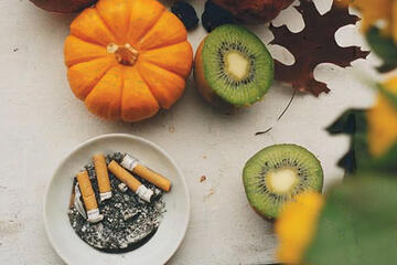A overhead photo of kiwis, mini pumpkin, and an ash tray filled with ash and four cigarettes.