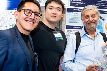 From left, Victor Ticllacuri, Yuxuan Bai, and Nitish Thakor pose in front of a poster display for their invention at WSE Design Day 2025. Nitish Thakor is holding a lifelike baby doll, used to demonstrate their invention