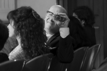 Black-and-white film still that shows a man sitting next to a woman in an auditorium but looking at the viewer with an eyeglass