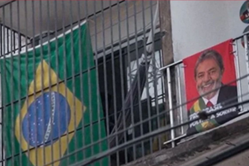 Film still from "No Céu da Pátria Nesse Instante" featuring a Brazilian flag, a balcony, and a portrait of a politician