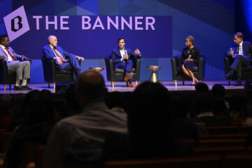 A panel of five individuals sits on stage; A sign reading The Banner is visible in the background