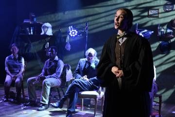 An actor in 1800s male clothing speaks while standing in a spotlight. Several actors in similar period attire sit on chairs in the background, draped in moody blue and green lighting.