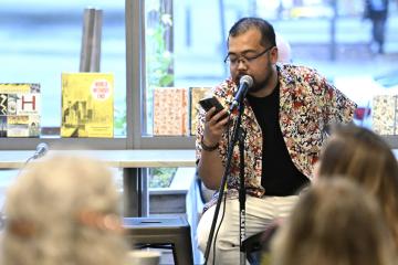 An adult reads off their phone into a microphone. People listen in front of them.