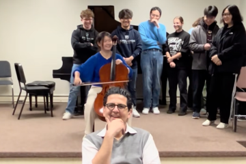 Amit Peled strokes his chin as a student plays the cello behind him. Behind the student, more classmates stand and listen.