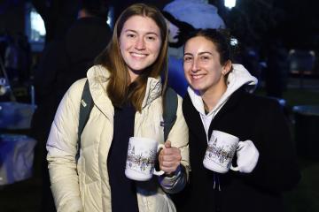 Two college students smile for a picture while holding up Lighting of the Quads 2025 mugs.