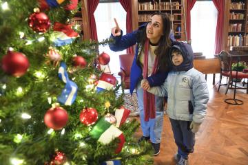 A parent points to the top of the Christmas tree. Their young child follows with their eyes.