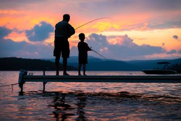 A grandfather and his grandchild fishing off a dock are silhouetted against a colorful sunset.