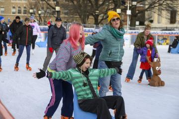 Three people skate on a public rink, surrounded by others. One is pushing another on a sled.