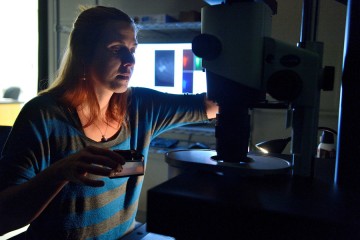 A woman holds a square of jelly in one hand and adjusts a knob on a microscope with the other hand