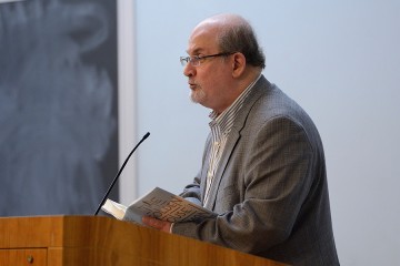 Salman Rushdie, dressed in a gray suit and striped shirt, holds open a hard cover copy of his book, The Golden House, and speaks into a podium microphone