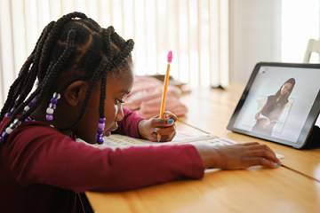 A child does classwork while sitting in front of a tablet displaying a video conference call