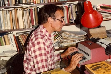 A man with glasses sits at a typewriter in a room with full bookshelves lining the walls