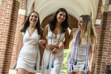Three young women in white dresses with Commencement stoles around their shoulders  link arm and walk toward the camera